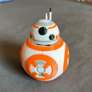 Może przedstawiać: Figurka droida BB-8 wydrukowana w 3D. Sferyczny korpus jest biały z pomarańczowymi panelami i akcentami. Głowa jest biała z szarymi i czarnymi detalami oraz dwiema antenami. Droid ma okrągły pomarańczowy wzór z przodu.