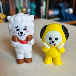 May include: Two small, colorful figurines. One is a white sheep-like character with brown arms and a red sash. The other is a yellow character with black ears and a white face. Both have cartoonish features.