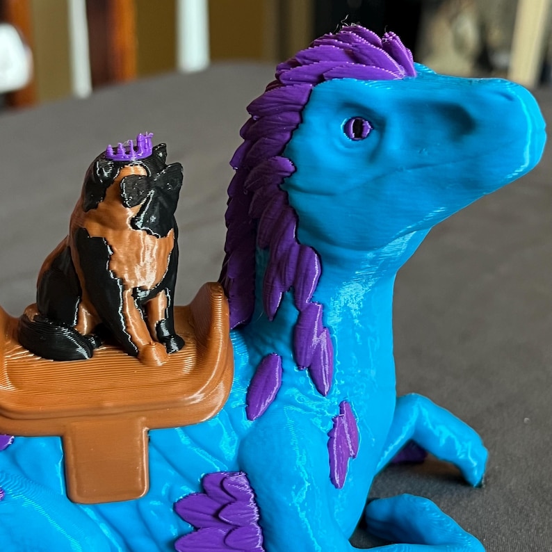 Puede incluir: Una criatura de fantas&iacute;a impresa en 3D con cuerpo azul, melena y detalles morados, y una peque&ntilde;a figura de perro con corona montada en su espalda. El perro es negro, marr&oacute;n y blanco. La silla es marr&oacute;n.