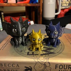 May include: Three small dragon figurines in black, gold, and navy blue. The dragons have large eyes and detailed wings. They are sitting on a book with gold lettering. The black dragon is on the left, the gold in the middle, and the navy blue on the right.