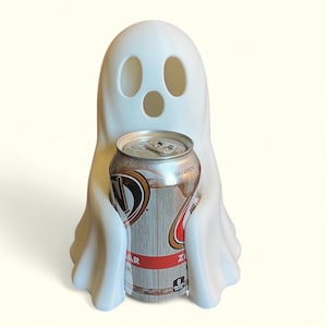 May include: A white ghost-shaped can cooler holding a silver and brown soda can. The ghost has three eye holes and a mouth hole. The soda can has red and brown text and says "No Caffeine".