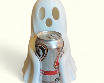 Puede incluir: Un enfriador de latas en forma de fantasma blanco que sostiene una lata de refresco plateada y marrón. El fantasma tiene tres agujeros para los ojos y un agujero para la boca. La lata de refresco tiene texto rojo y marrón y dice "Sin cafeína".