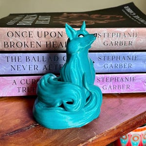 May include: A teal blue 3D printed figurine of a fox with a curled tail. The fox is sitting on a stack of books with the author Stephanie Garber's name on the spines.