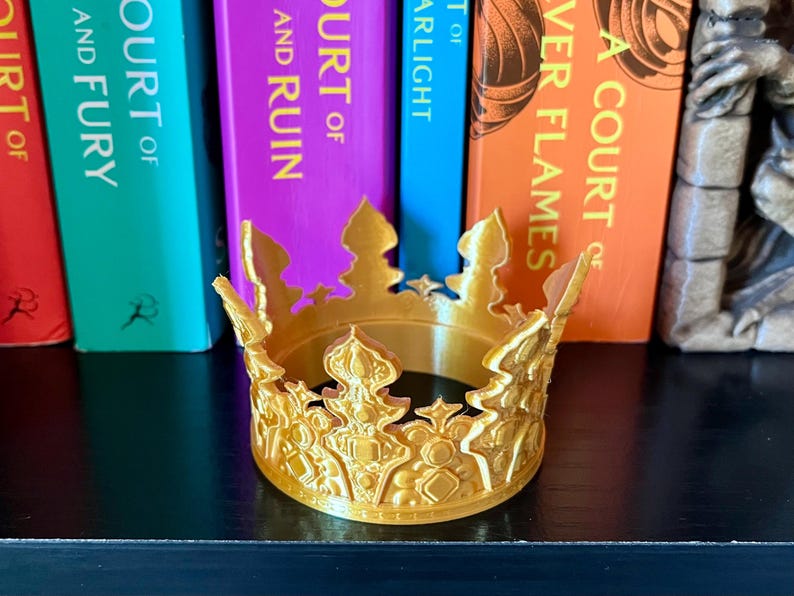 May include: A gold-colored crown with intricate details, sitting in front of a row of colorful books. The crown has a circular base and ornate embellishments. The books have titles like "A Court of Thorns and Roses" and "A Court of Silver Flames."