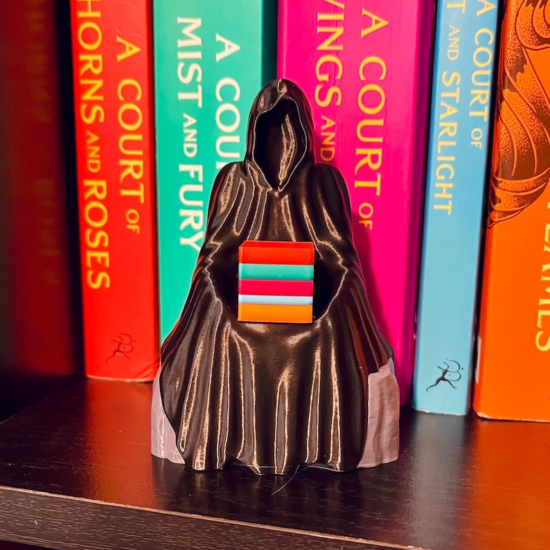 Op de afbeelding: Een zwart beeldje van een figuur met een capuchon die een stapel kleurrijke rechthoekige blokken vasthoudt. Het beeldje staat op een plank voor een rij boeken met titels als "A Court of Thorns and Roses".