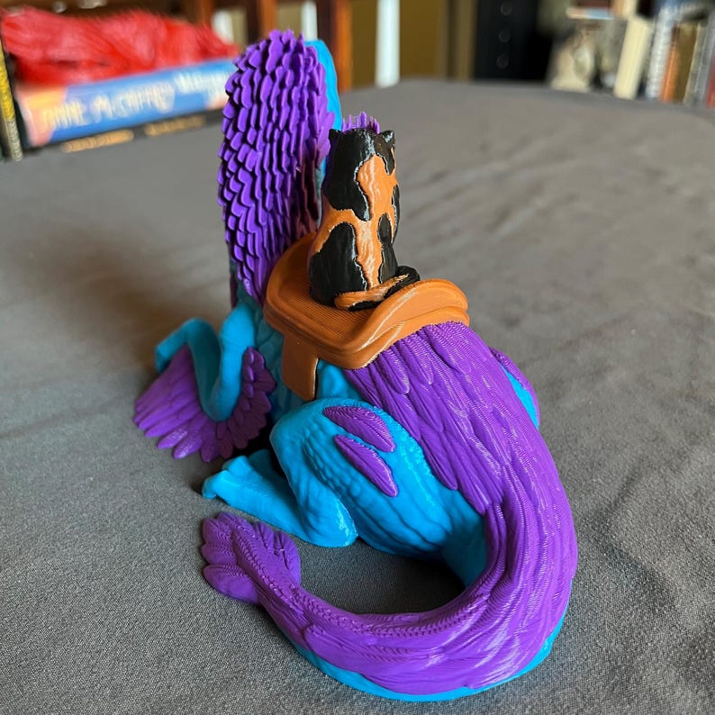 Puede incluir: Una figura de fantas&iacute;a de una criatura alada en tonos morados y azules, con un peque&ntilde;o gato posado en su espalda. El gato es negro y naranja, y la figura tiene una silla de montar. La figura es un art&iacute;culo decorativo &uacute;nico.