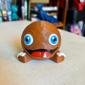 Puede incluir: Una figura de criatura esf&eacute;rica marr&oacute;n impresa en 3D con grandes ojos azules, una boca negra ancha, una lengua roja y un solo diente blanco. El personaje tiene peque&ntilde;as manos con puntas blancas y un cuerpo liso y redondeado.