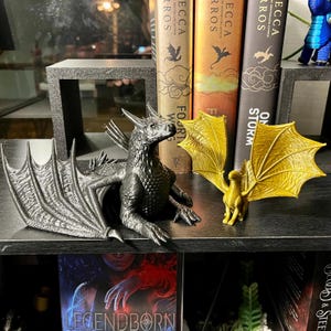 May include: Two dragon figurines, one black and one gold, are displayed on a shelf. The black dragon has detailed wings and a textured body. The gold dragon has spread wings. Books with dragon illustrations are in the background.