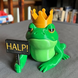 Puede incluir: Una figura de rana verde con una corona dorada que sostiene un letrero negro que dice "HALP!". La rana tiene una expresión ligeramente gruñona y está sentada sobre una superficie gris. Es un objeto decorativo caprichoso.