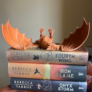 May include: An orange dragon figurine rests atop a stack of three books. The books are titled "Fourth Wing", "Iron Flame", and "Onyx Storm" by Rebecca Yarros. The dragon has outstretched wings and is lying on its back.