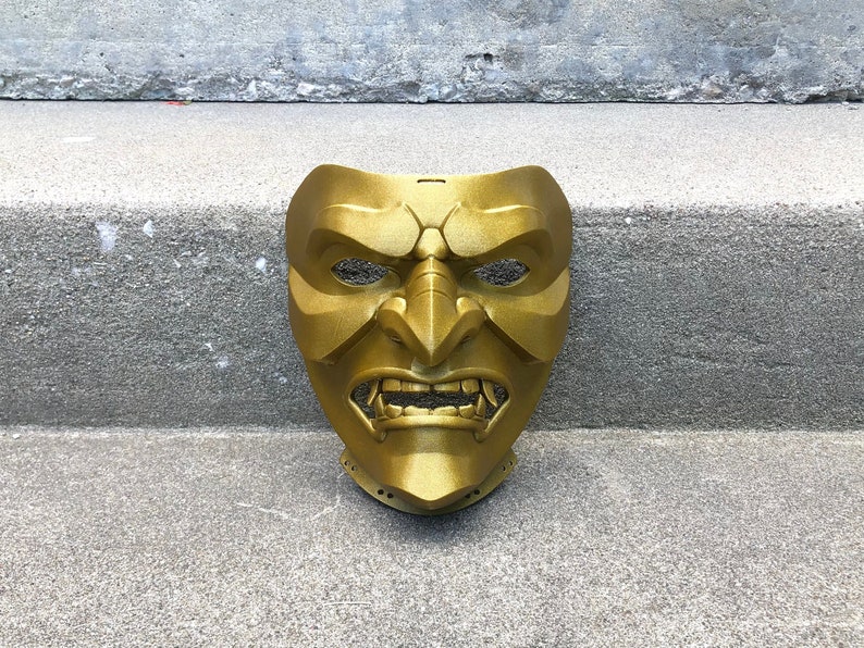 May include: A gold-coloured, plastic Oni mask with a fierce expression. The mask has a wide mouth with sharp teeth, and the eyes are narrowed and intense.