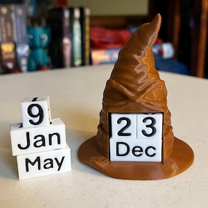 以下が含まれることがあります： 茶色の3Dプリントされた、組み分け帽子型の万年カレンダー。帽子には日付と月を表示する白い立方体が入っています。表示されている日付は12月23日です。