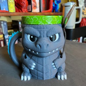 May include: A gray Godzilla-themed mug with a lime green rim and a blue tail. The mug has a handle and a silver spoon is placed inside. The mug is decorated with a Godzilla design.