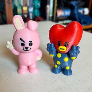 May include: Two 3D-printed figurines. One is a pink bunny with a white paw raised. The other is a blue figure with yellow polka dots and a red heart-shaped head. Both have black eyes and are standing.