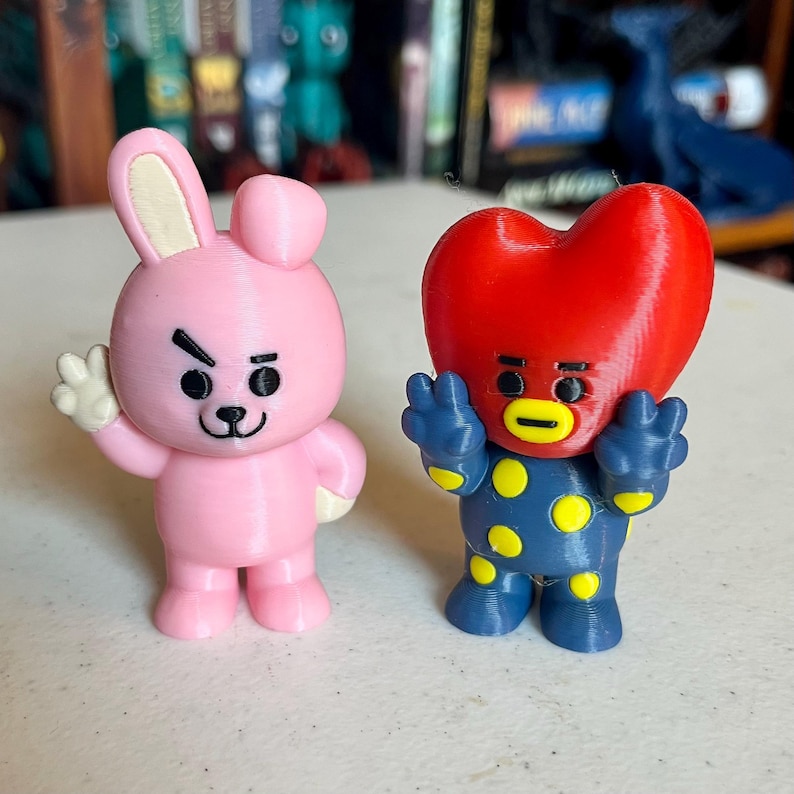 May include: Two 3D-printed figurines. One is a pink bunny with a white paw raised. The other is a blue figure with yellow polka dots and a red heart-shaped head. Both have black eyes and are standing.