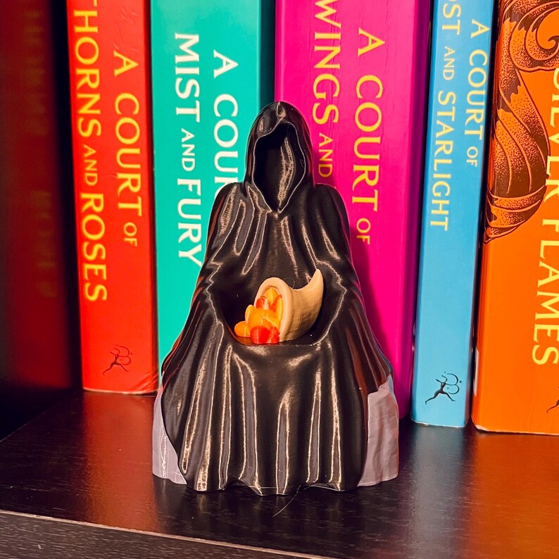Op de afbeelding: Een zwarte figuur met capuchon die een hoorn des overvloeds vasthoudt, gevuld met kleurrijk fruit en groenten. Het beeldje staat voor een rij kleurrijke boeken met titels als "A Court of Thorns and Roses" en "Silver Flames".