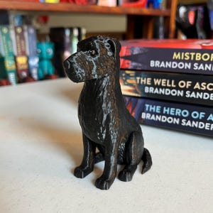 以下が含まれることがあります： 座った姿勢の黒い3Dプリント犬の置物。犬はテクスチャ加工された表面を持ち、「ミストボーン」、「アセンションの井戸」、およびブランドン・サンダーソンの「エイジズの英雄」というタイトルの本のスタックの前に配置されています。