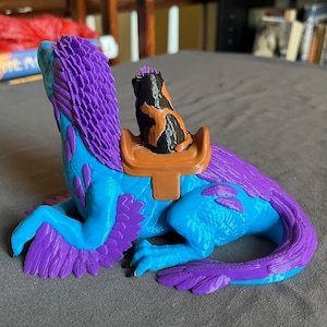 Puede incluir: Una figura de criatura fant&aacute;stica impresa en 3D en tonos azules y morados, con una silla de montar que sostiene una figura de gato. La criatura tiene una larga cola y alas, y el gato es negro y naranja. La figura est&aacute; sobre una superficie gris.
