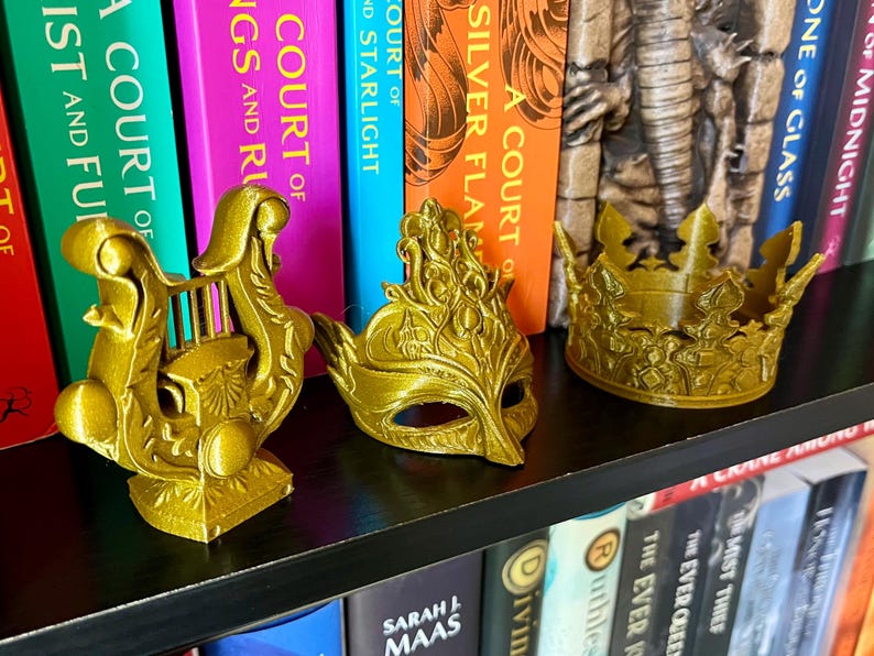 May include: Three gold-colored decorative items: a lyre, a masquerade mask, and a crown. The items are displayed on a black shelf in front of a row of colorful books. The books have titles like "A Court of Silver Flames" and "A Court of Thorns and Roses."