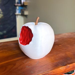 Puede incluir: Escultura de cer&aacute;mica blanca con forma de manzana a la que le falta un mordisco, revelando un interior rojo texturizado. La manzana tiene un tallo marr&oacute;n y est&aacute; sobre una superficie de madera. Es un objeto decorativo.