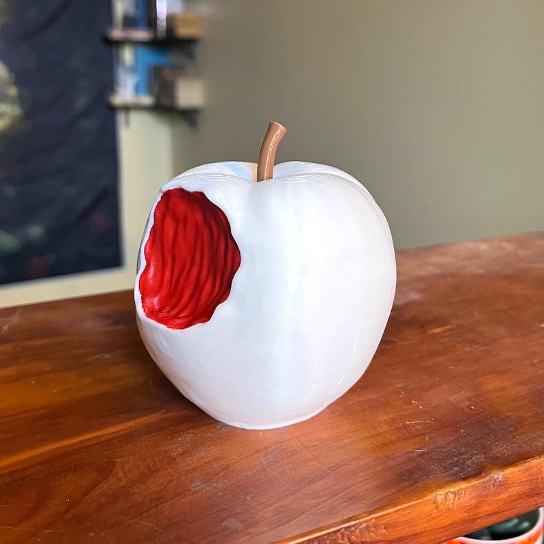 Puede incluir: Escultura de cer&aacute;mica blanca con forma de manzana a la que le falta un mordisco, revelando un interior rojo texturizado. La manzana tiene un tallo marr&oacute;n y est&aacute; sobre una superficie de madera. Es un objeto decorativo.