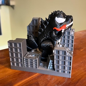 May include: A black Godzilla figure with a Venom-inspired design, featuring a white eye and red tongue, stands amidst gray miniature city buildings. The figure is a unique blend of two iconic characters.