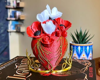 Op de afbeelding: Een 3D-geprint rood hart met gouden ranken, dat een boeket witte en rode bloemen vasthoudt. Het hart staat op een gouden standaard en ligt op een zwart boek met de titel "Lightbringer" van Alex Aster.
