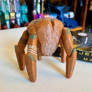 May include: A brown, articulated crab-like figurine with green accents on its legs. The figurine has a textured body and detailed legs with etched designs and white stripes. It stands on four legs, showcasing a unique, artistic design.