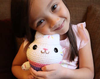 Puede incluir: Un peluche de conejo blanco hecho a mano con ganchillo, con ojos negros, una nariz rosa y una parte inferior a rayas de colores. El conejo está siendo sostenido por una niña pequeña que lleva una camisa rosa y leggings grises con estrellas doradas.