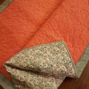 May include: A coral-colored quilted throw blanket with a floral pattern on the reverse side. The blanket has a gray border.