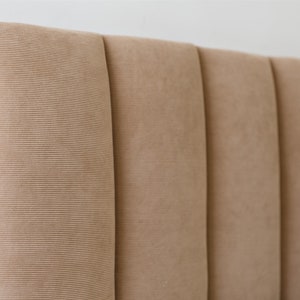 May include: A close-up of a brown headboard with vertical channel tufting. The headboard is made of a soft, textured fabric.