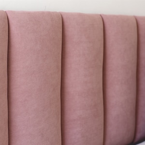May include: A close-up of a pink headboard with vertical tufted panels. The headboard is made of a soft, textured fabric.