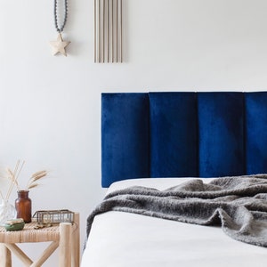 May include: A navy blue upholstered headboard with vertical paneling is the focal point of this bedroom scene. A gray blanket is draped over the white bed linens. A small side table holds decorative items, including a vase with wheat stalks.