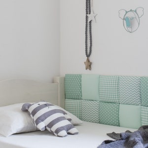 May include: A white bed with a green and white patterned headboard. The headboard is made of individual squares of fabric with different patterns. There is a white pillow and a gray and white striped stuffed animal on the bed.