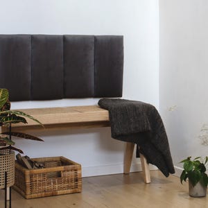May include: A wooden bench with a grey upholstered headboard and a grey throw blanket. The bench is in a room with white walls and a wooden floor. There are plants in pots on either side of the bench.