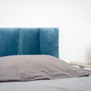 K&ouml;nnte beinhalten: Ein blaues, gepolstertes Kopfteil mit einem grauen Kissen auf einem Bett mit einer grauen Bettdecke. Ein kleiner Beistelltisch mit einem Buch und einer Topfpflanze ist im Hintergrund zu sehen.