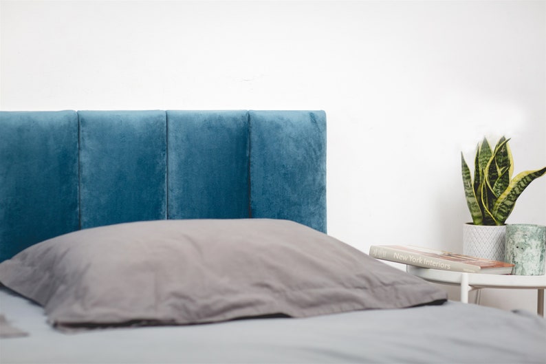 K&ouml;nnte beinhalten: Ein blaues, gepolstertes Kopfteil mit einem grauen Kissen auf einem Bett mit einer grauen Bettdecke. Ein kleiner Beistelltisch mit einem Buch und einer Topfpflanze ist im Hintergrund zu sehen.