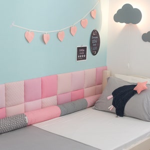 Op de afbeelding: Een lichtblauwe muur met een roze hartengirlande, een grijze wolkvormige wandlamp en een wit bed met een roze en grijze gecapitonneerde hoofdeinde. Het bed heeft een grijs-wit gestreept kussen en een roze-wit sterrenkussen. De muur heeft ook een zwart krijtbord met de tekst "Dream Big Little One" en een wit bord met de tekst "Little Rock" en vijf sterren.