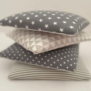 May include: A stack of four decorative throw pillows with different geometric patterns. The top pillow has white stars on a gray background, the second pillow has a geometric pattern in beige and white, the third pillow has white polka dots on a gray background, and the bottom pillow has white stripes on a beige background.