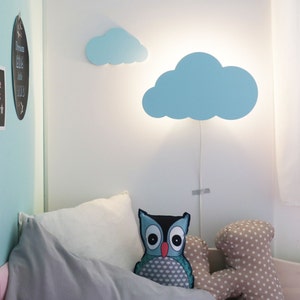 May include: A light blue cloud-shaped wall lamp with a white cord hanging from it. The lamp is mounted on a white wall. There is a bed with gray and white bedding and a blue and gray owl pillow in the foreground.