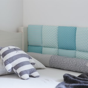 May include: A white bed with a gray and white striped pillow and a gray and white stuffed animal. The headboard is made of a patchwork of blue and green fabric with different patterns.