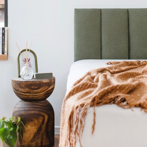 May include: A wooden bedside table with a brown finish and a round top. The table has a unique hourglass shape and is decorated with a clear glass vase with dried reeds and a small rectangular speaker. A green headboard with vertical panels is in the background.