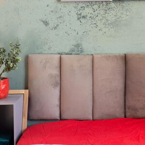 May include: A bedroom scene featuring a headboard with five vertical panels in a muted taupe color. A vibrant red blanket is visible at the foot of the bed. A red geometric pot with a green plant sits on a side table.