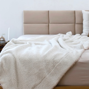 May include: A white fluffy blanket draped over a bed with a beige headboard. The bed is made with white sheets and a brown pillow.