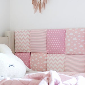 Puede incluir: Una cabecera acolchada rosa y blanca con una variedad de patrones, incluyendo chevron, rayas y nubes. Un conejito de peluche blanco se sienta sobre una manta rosa frente a la cabecera.