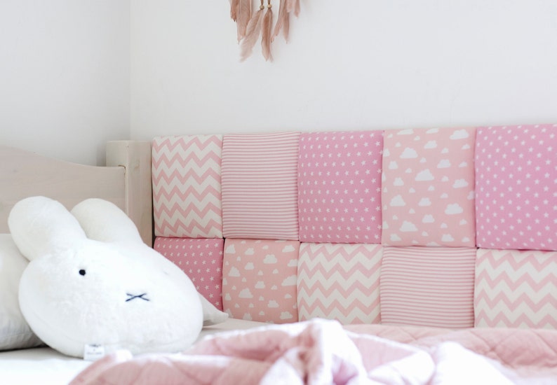 Puede incluir: Una cabecera acolchada rosa y blanca con una variedad de patrones, incluyendo chevron, rayas y nubes. Un conejito de peluche blanco se sienta sobre una manta rosa frente a la cabecera.