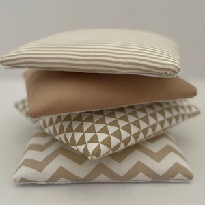 Op de afbeelding: Een stapel van vier decoratieve kussens met verschillende geometrische patronen in beige en wit. Het bovenste kussen heeft horizontale strepen, het tweede kussen is effen beige, het derde kussen heeft een geometrisch patroon van driehoeken en het onderste kussen heeft een chevron-patroon.