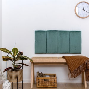 May include: A teal upholstered headboard with three panels, a wooden bench with a woven seat, a brown throw blanket, a potted plant, and a clock on a white wall.