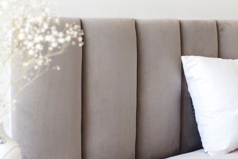 May include: A close-up of a grey upholstered headboard with vertical channel tufting. The headboard is part of a bed with a white pillow on top.