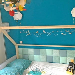 Puede incluir: Una cama con estructura de madera y cabecero acolchado. La pared es de color azul turquesa. El cabecero est&aacute; hecho de un mosaico de cuadrados de tela azul claro y gris. Un m&oacute;vil de nube blanca cuelga del techo.